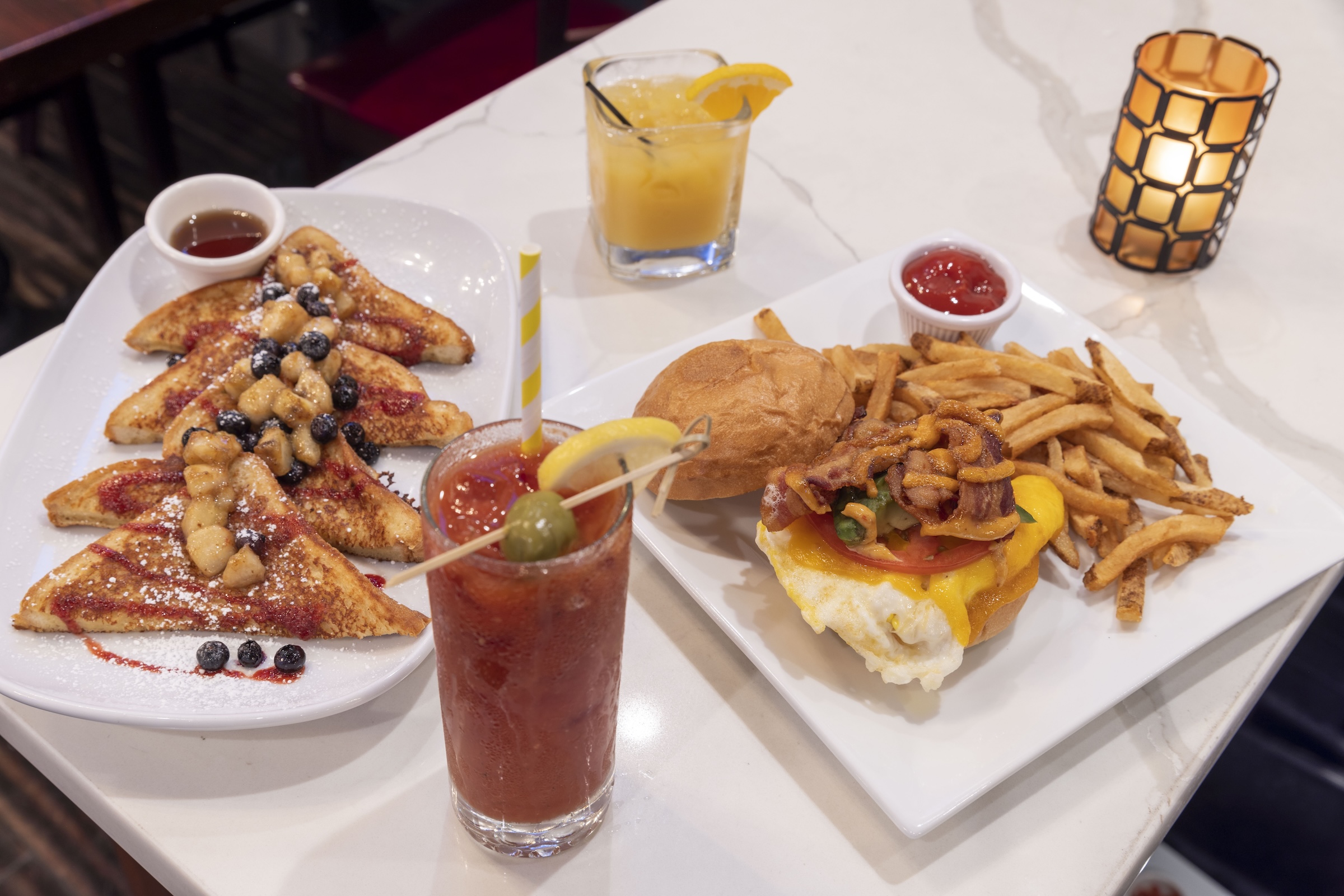 A high-angle close-up of a brunch spread on a white marble tabletop. On the left, a plate of French toast triangles is topped with powdered sugar, berries, and caramelized bananas, served with a small side of syrup. To the right, a breakfast burger on a brioche bun features a fried egg, melted cheese, and bacon, served with a large portion of french fries and ketchup. In the foreground, there is a tall Bloody Mary garnished with a lemon slice and olive, alongside a glass of orange juice and a decorative candle holder in the background.
