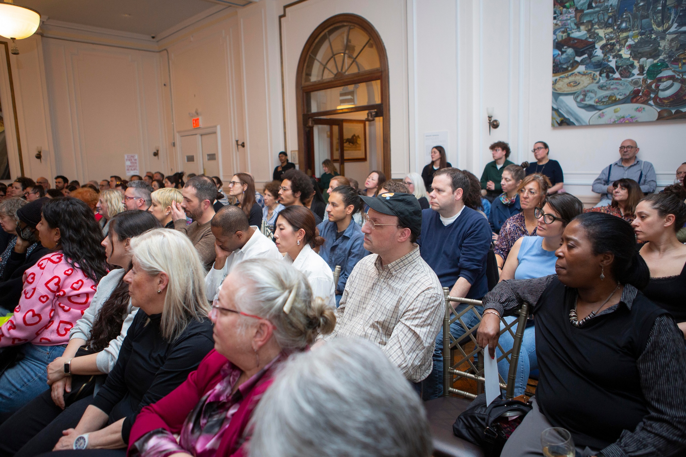 A high-angle shot of a crowded room filled with people of various ages and backgrounds sitting in rows of gold-colored chairs. The audience is looking toward a stage (not pictured). The setting is a formal hall with high ceilings, cream-colored walls, large arched doorways, and a colorful piece of modern art hanging on the right wall. The atmosphere appears focused and attentive.