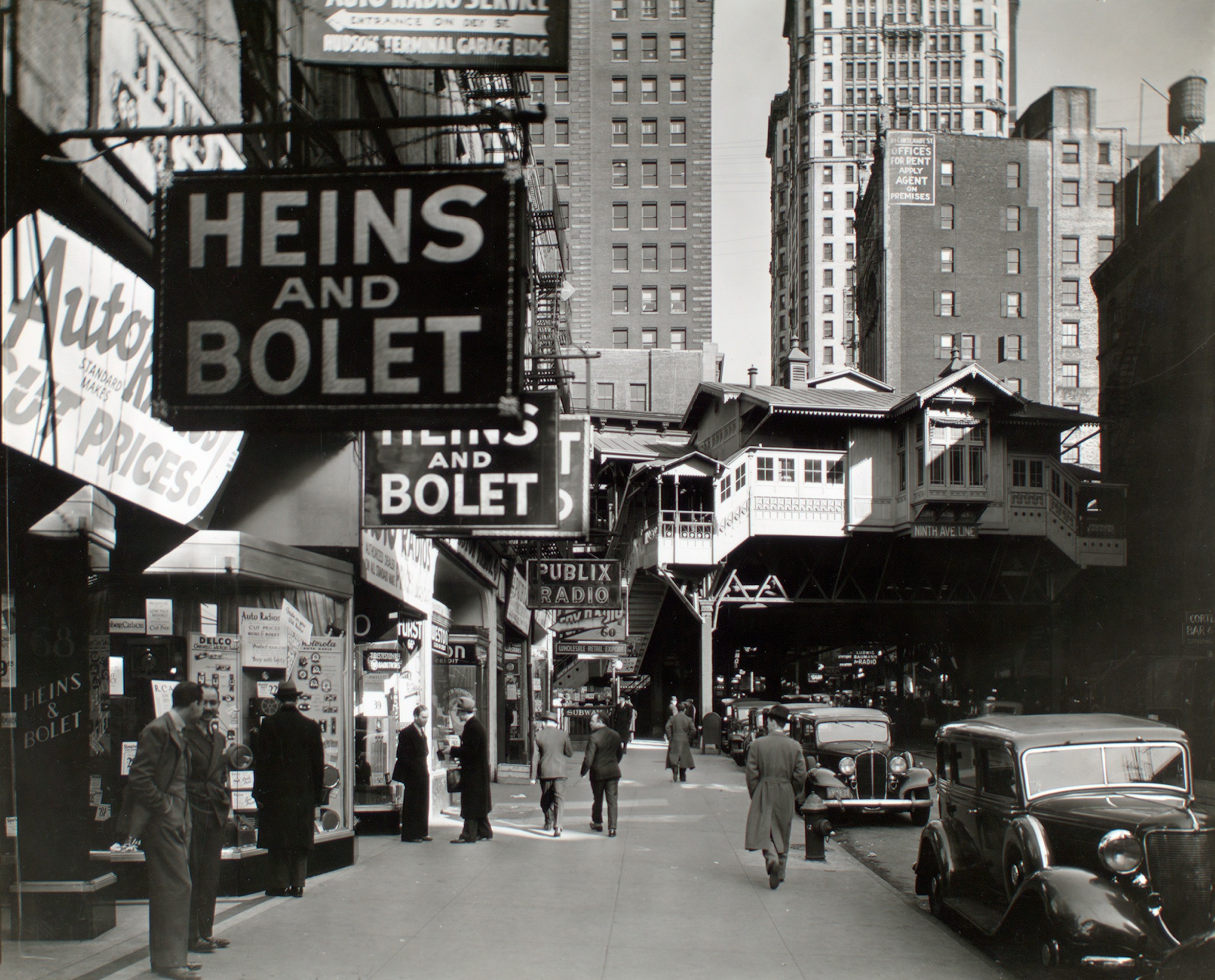 A black-and-white historical photograph of Cortlandt Street in New York City, known as Radio Row. Large hanging signs for "Heins and Bolet" and "Publix Radio" line the sidewalk where men in overcoats and hats walk past storefronts. In the background, an elevated train station for the Ninth Avenue Line spans across the street, framed by towering Manhattan skyscrapers.