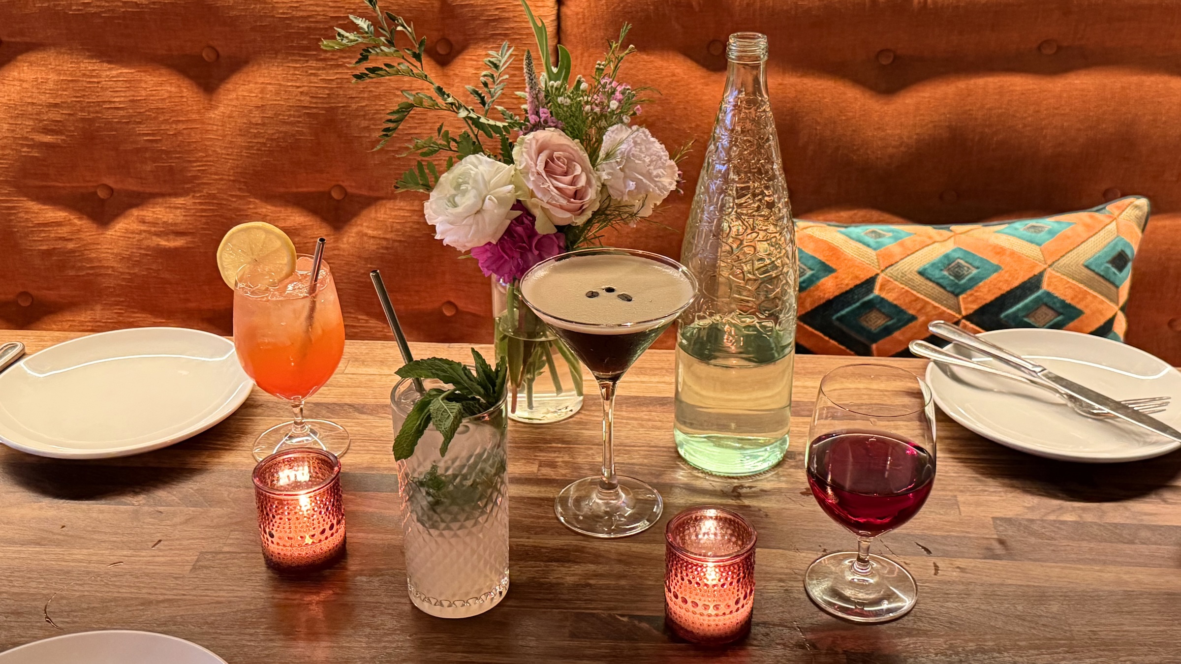 A warm, eye-level shot of a wooden restaurant table set with various colorful cocktails and drinks. In the center sits an espresso martini in a coupe glass, flanked by a tall mojito with fresh mint leaves and a vibrant orange spritz garnished with a lemon wheel. A glass of red wine sits to the right next to a textured clear glass water bottle. Two small pink glass votive candles cast a warm, flickering glow on the table. In the background, a small floral arrangement with pink and white roses rests against an orange button-tufted banquette with a geometric patterned pillow.