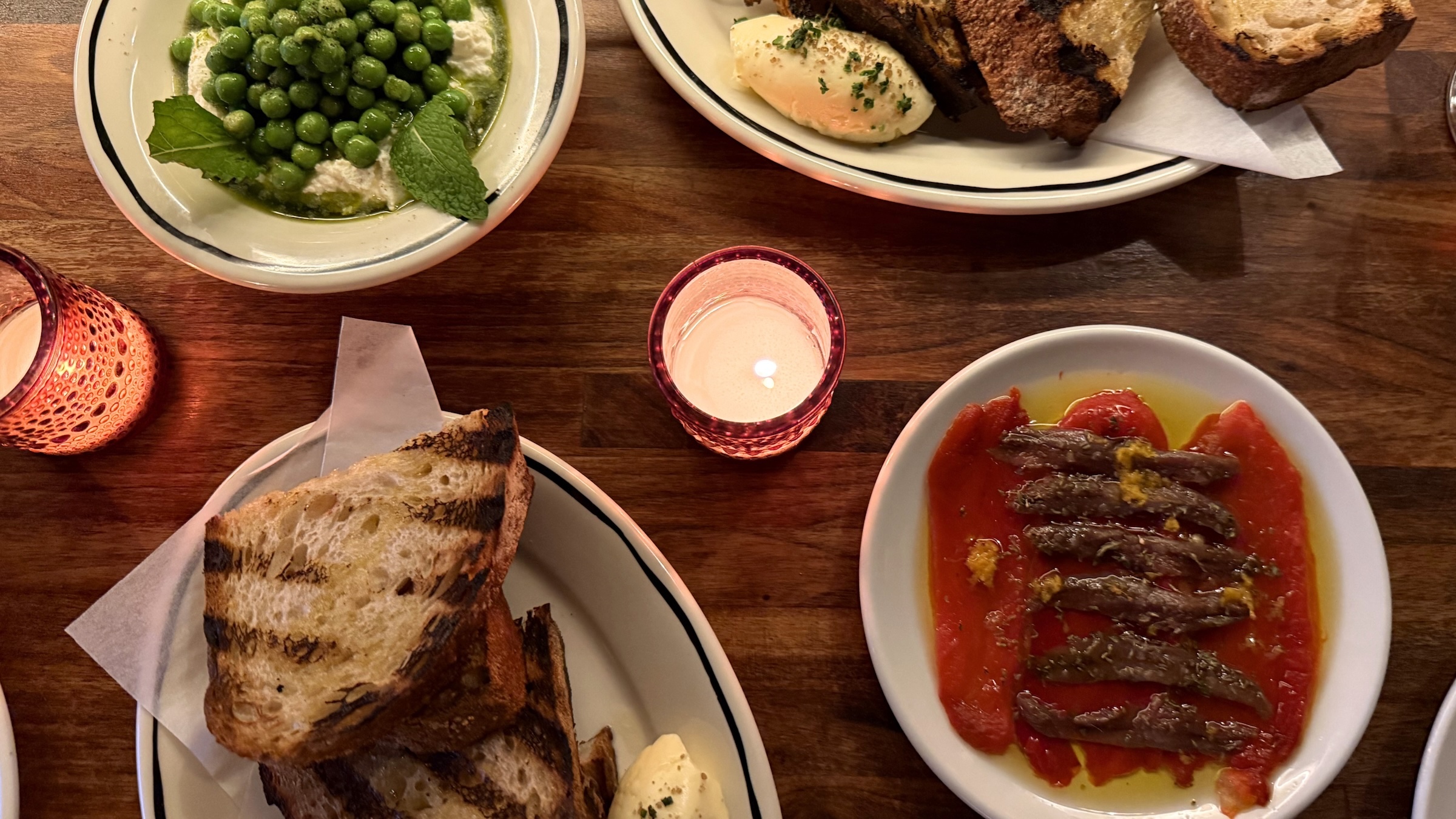 A top-down view of an assortment of appetizers served on a dark wooden table, illuminated by a central pink glass votive candle. On the bottom right, a small white plate holds silver anchovy fillets laid over a bright red pepper base, drizzled with olive oil and herbs. Two plates feature thick, char-grilled slices of rustic sourdough bread served with a dollop of whipped butter. In the upper left, a shallow white bowl contains bright green English peas resting on a bed of white ricotta or burrata, garnished with fresh mint.