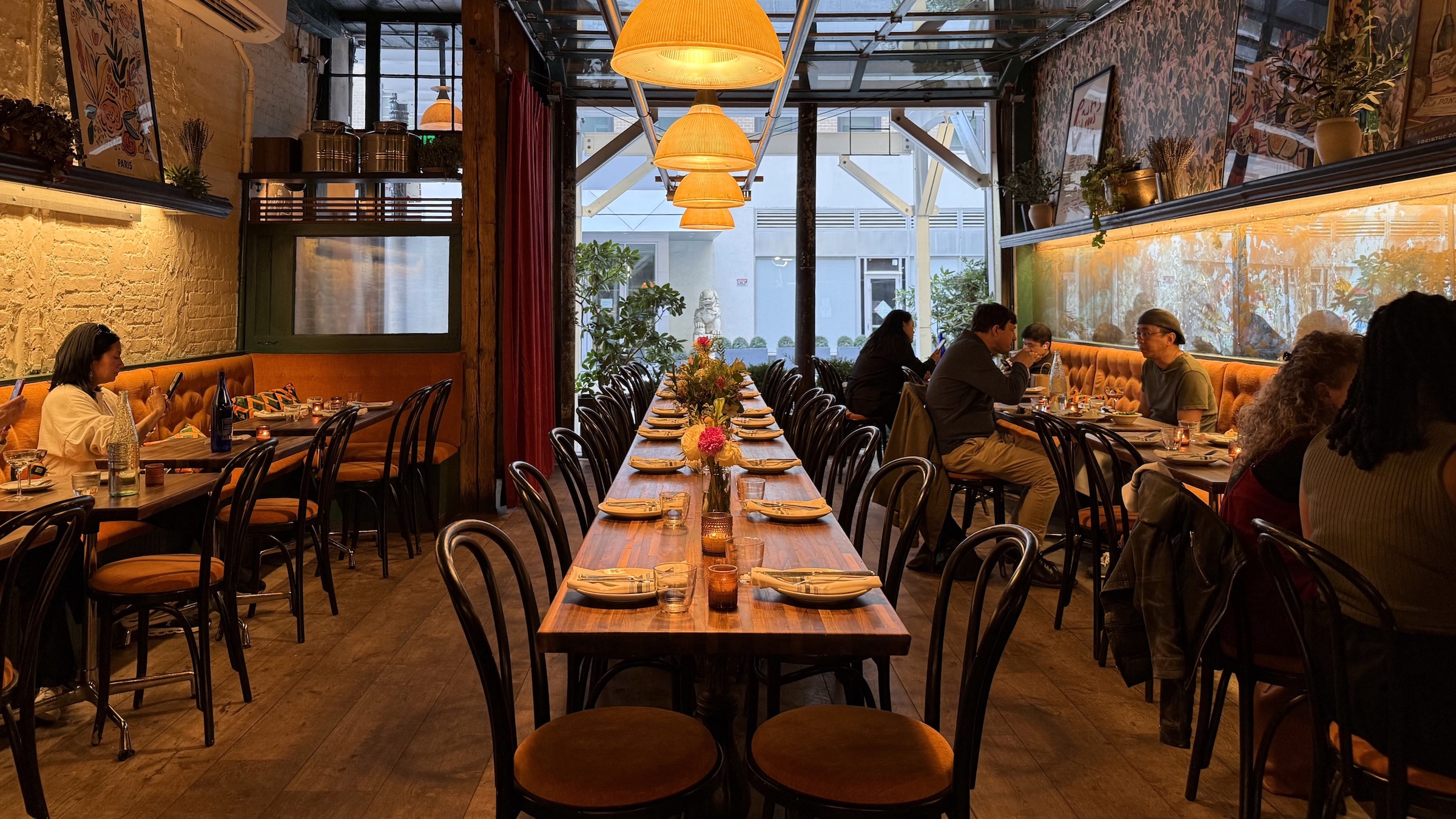 A wide interior shot of a narrow, cozy restaurant with a rustic yet refined aesthetic. A long, wooden communal table fully set with plates, silverware, and glassware stretches down the center under a row of low-hanging, warm yellow dome pendant lights. Patrons sit at smaller tables along the sides on bentwood chairs with orange upholstered seats. The left wall features exposed brick and a tufted orange banquette, while the right wall is adorned with patterned wallpaper. The back of the restaurant opens up into a bright atrium space through a glass garage-door style ceiling structure.