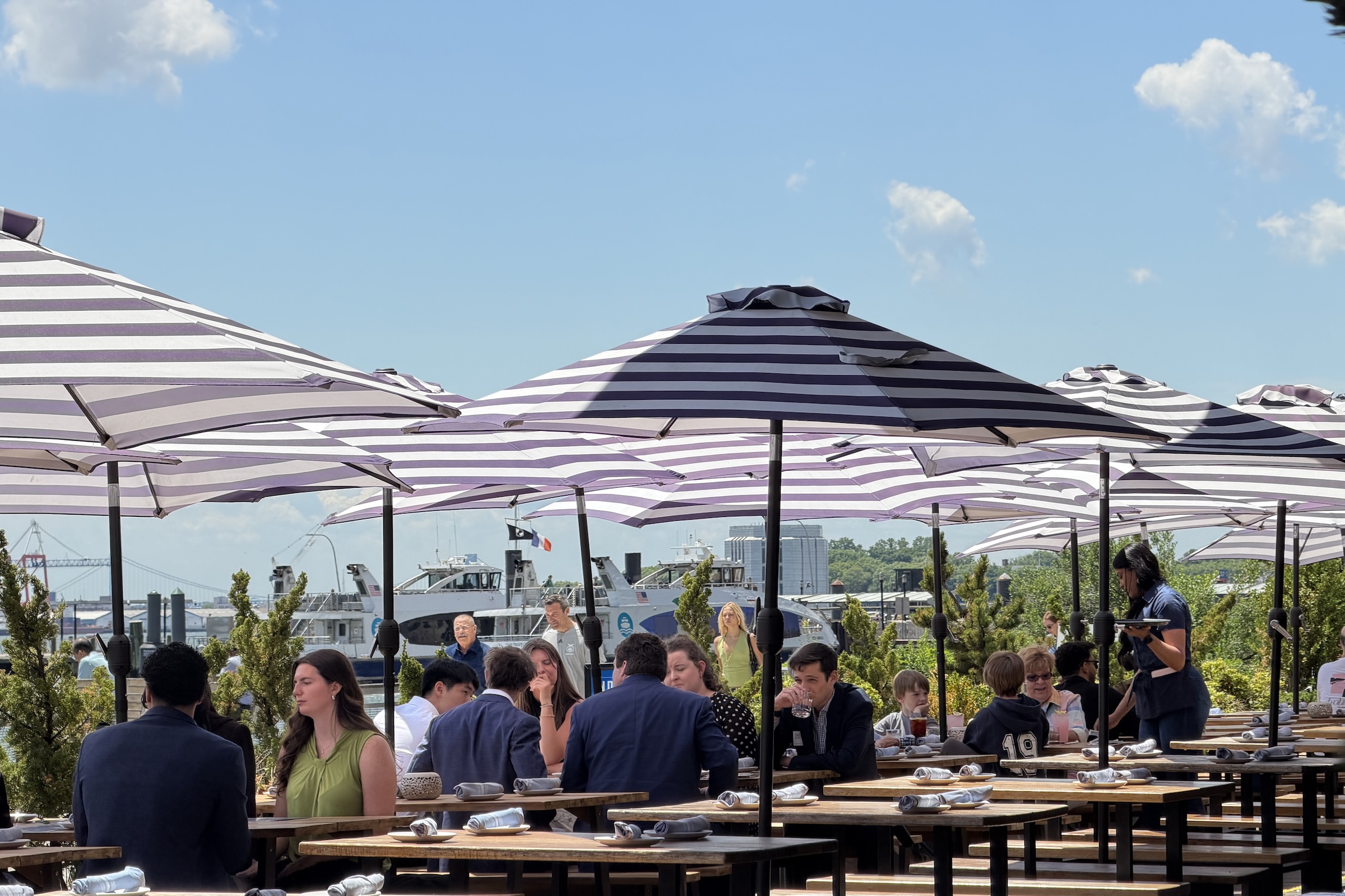 A daytime view of a bustling outdoor restaurant patio situated on a waterfront. Multiple long wooden tables are packed with patrons talking and dining. Overhead, an array of wide, black-and-white striped patio umbrellas provides shade. A server stands to the right holding a tray. In the background, large white passenger boats and ferries are docked along the water, with a distant bridge suspension tower visible under a clear blue sky with fluffy white clouds.