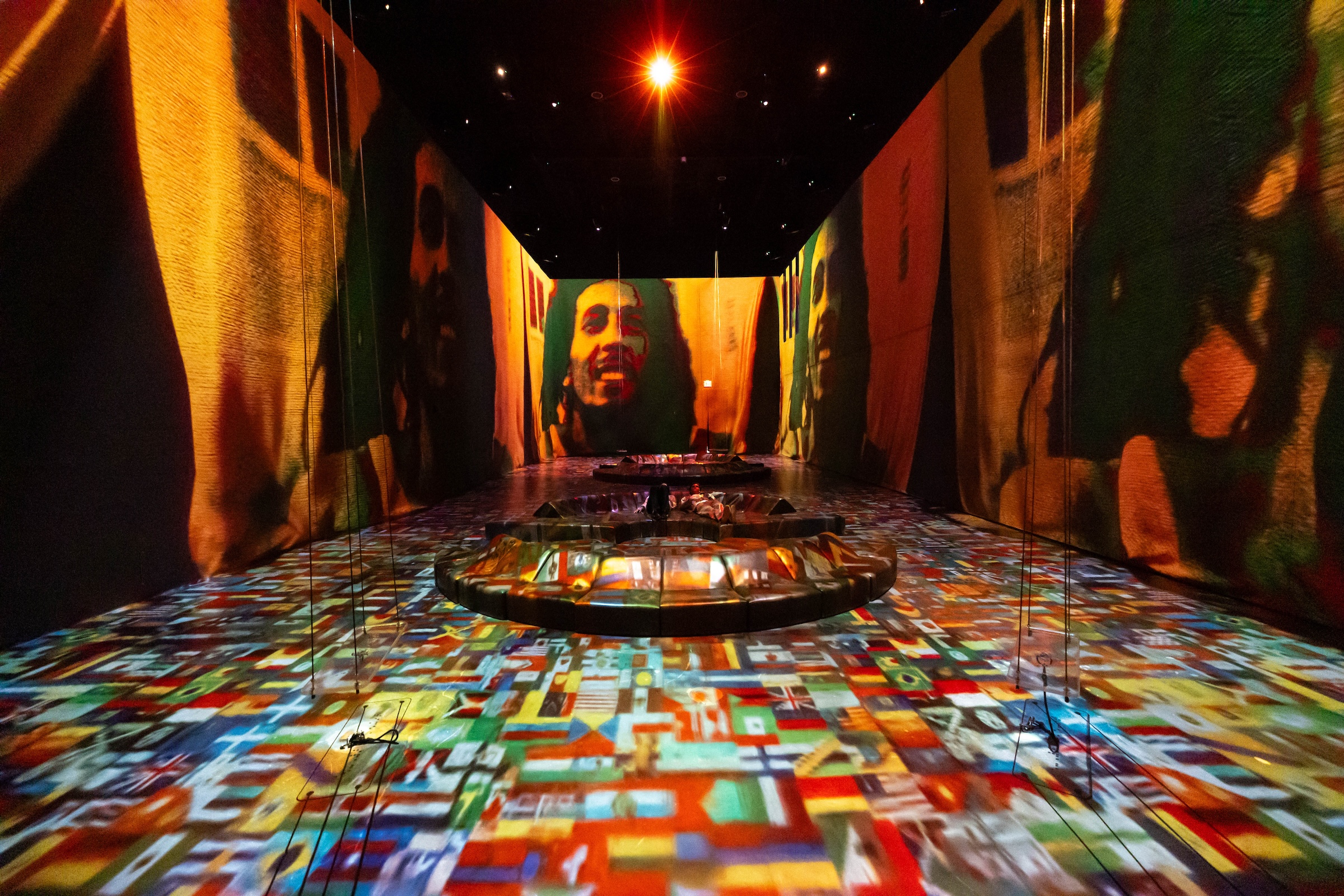 A wide-angle shot of an immersive exhibition room with black walls and ceilings. High-contrast, stylized portraits of Bob Marley are projected onto the large fabric walls. The floor is vibrant, entirely covered by a projection of numerous international flags. A bright, lens-flare spotlight shines from the center of the ceiling over circular, dark seating structures in the middle of the room.