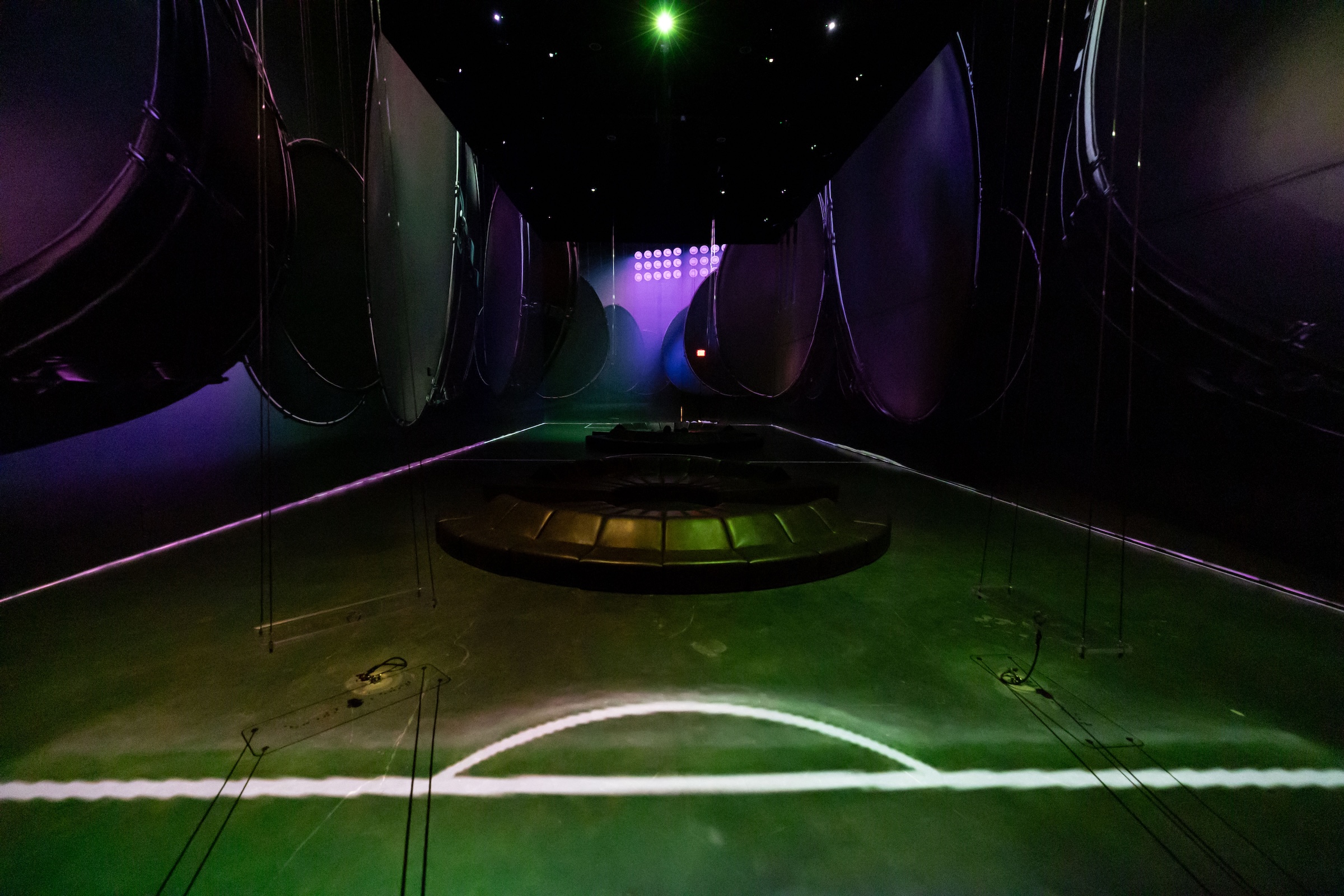 A dimly lit, immersive exhibition space with moody green and purple lighting. Large, circular metallic discs hang in rows along both sides of the ceiling. A green projection on the floor mimics the white center lines of a soccer field. A multi-tiered, circular dark bench sits empty in the center of the room under a single green light overhead.