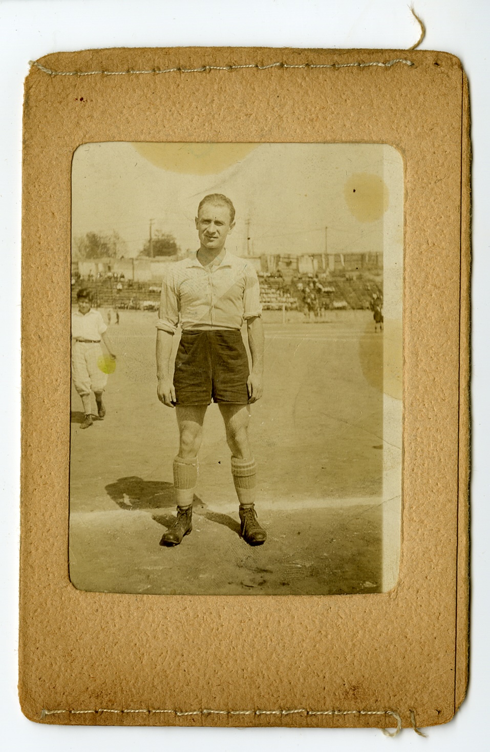 Vintage full-length portrait of an early 20th-century soccer player in a collared shirt, high-waisted shorts, and lace-up boots, standing on a field in front of stadium bleachers.