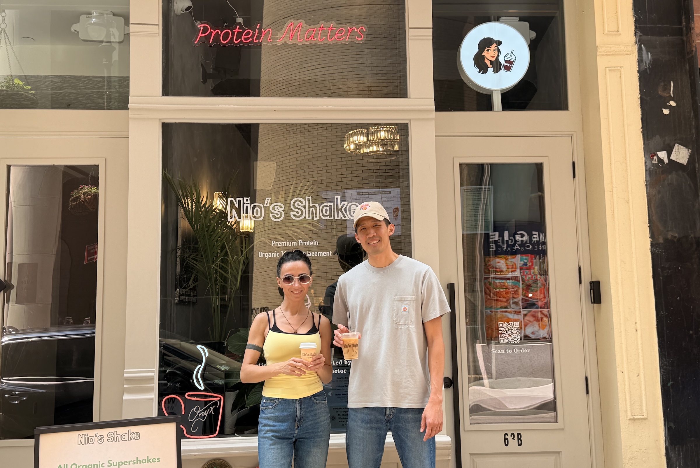 A full shot captures a man and a woman standing outside the storefront of a cafe named "Nio's Shake." The woman, on the left, has dark hair pulled back, sunglasses, and is wearing a yellow tank top and blue jeans while holding a small cup. The man, on the right, wears a light gray t-shirt, blue jeans, and a beige baseball cap, and holds a transparent plastic cup with an iced drink. Behind them, the shop's glass window displays white lettering reading "Nio's Shake" and "Premium Protein," with a pink neon sign above that reads "Protein Matters." A circular sign featuring a cartoon illustration of a woman holding a shake is mounted to the right of the entrance door.