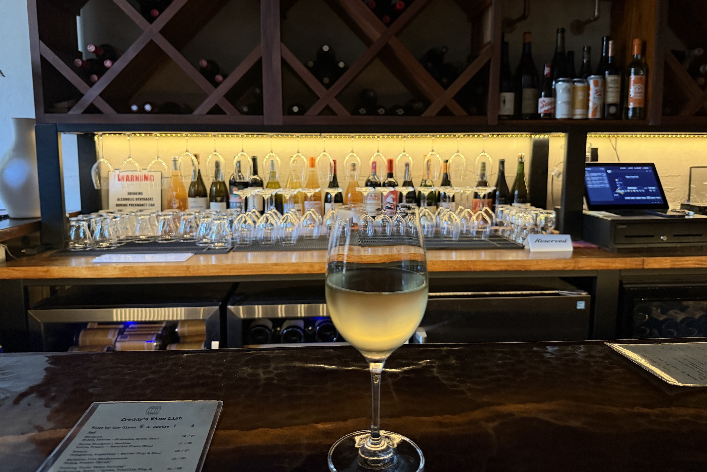 A close-up shot of a single glass of white wine resting on a dark, glossy, hammered copper bar counter. In the background, a warmly lit bar features rows of inverted wine glasses hanging under a shelf, various liquor and wine bottles, and a wooden wine rack filled with bottles. A menu sits on the counter to the left.