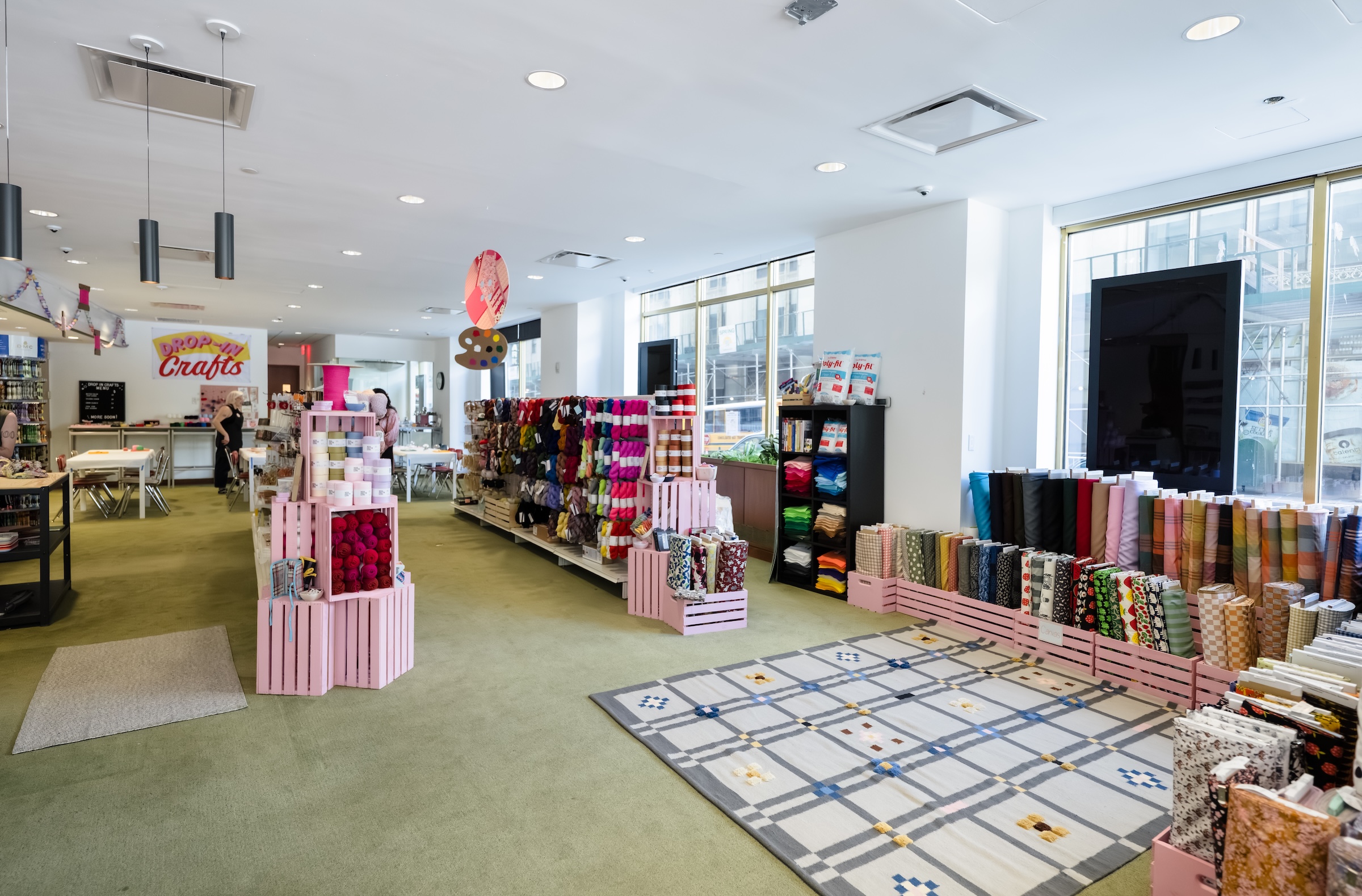 A brightly lit, bustling craft and fabric supply store with green carpeting and white walls. In the foreground, a large plaid area rug lies on the floor next to rows of colorful fabric bolts organized in pink wooden crates. In the center, stacks of pink crates display yarn, leading toward long shelves overflowing with multi-colored yarn skeins. In the far background, people gather around crafting tables underneath a sign that reads "DROP-IN Crafts."
