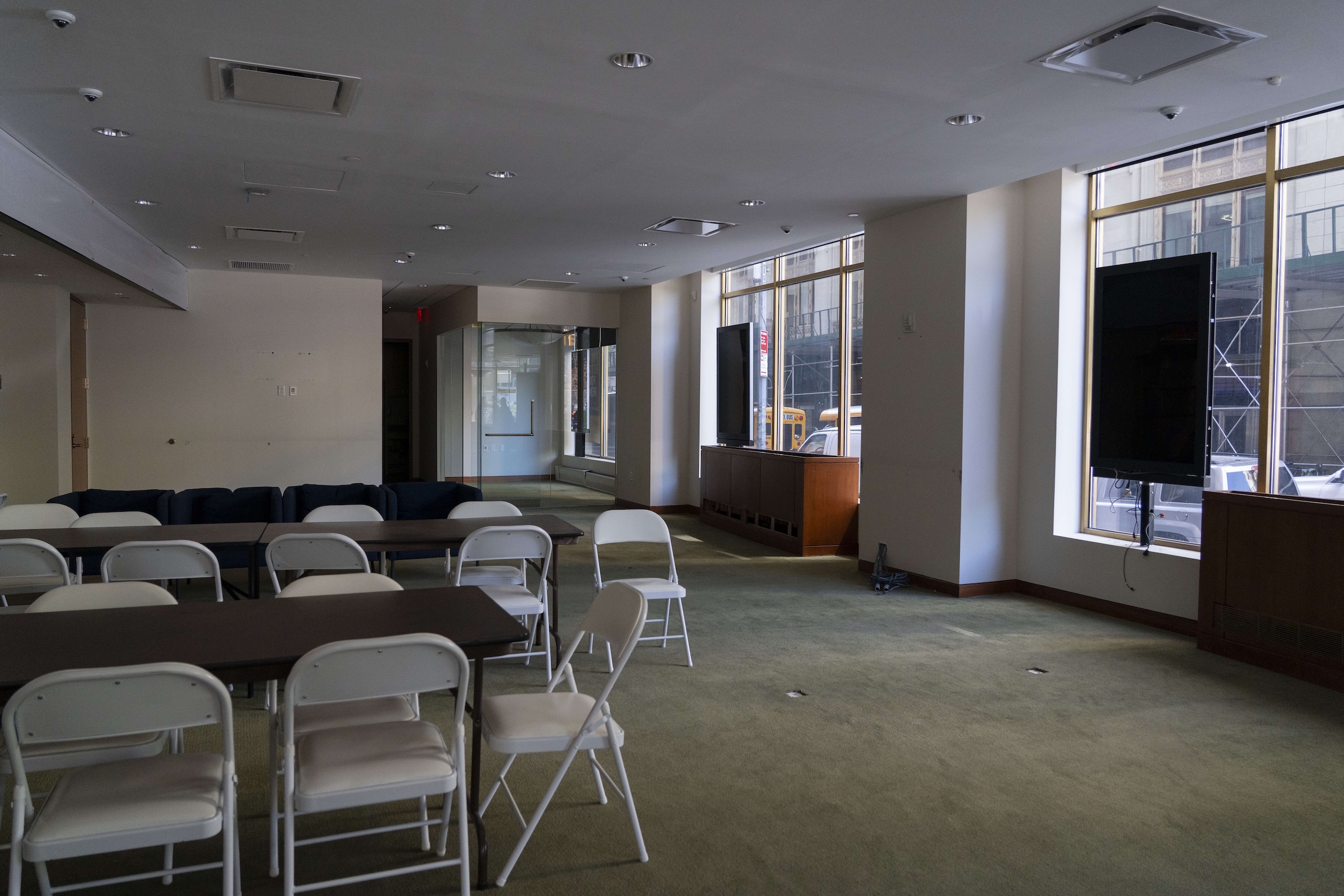 A dimly lit, sparse office or meeting room space with sage green carpeting and plain white walls. Several long brown folding tables are paired with rows of white plastic folding chairs, arranged in a classroom style. In the background, two large, black flat-screen monitors stand turned off on wooden consoles in front of large windows.