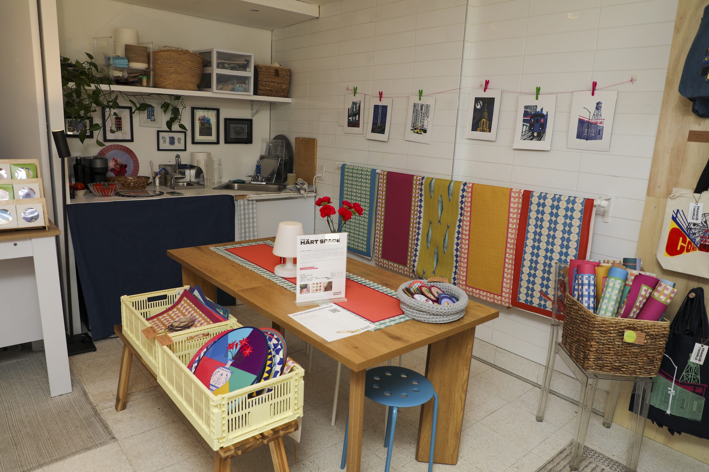 A brightly lit, vibrant boutique or community market space with white tiled walls and a light floor. In the center sits a wooden dining table draped with a red runner, a sign reading "THE HÄRT SPACE," red carnations, and a small blue stool underneath. Yellow plastic crates filled with colorful patterned plates rest on a bench nearby. A wide variety of brightly colored, patterned textiles hang neatly along the back wall, below a clothesline pinning up art prints. To the left, a small kitchenette area with a sink features shelves filled with woven baskets, plants, and framed pictures.