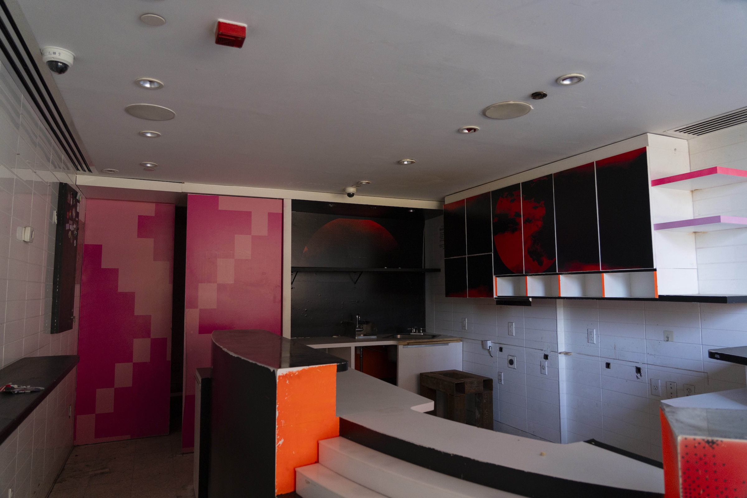 A dimly lit, vacant commercial kitchen or café interior undergoing a transition. The space features white tiled walls, a dark ceiling with security cameras, and an L-shaped service counter with bright orange trim. The back wall includes black kitchen cabinets adorned with a large red spherical graphic, next to a black-painted alcove over a sink. On the left side of the room, tall accent doors or panels are painted with a pixelated gradient pattern in shades of hot pink and light pink.