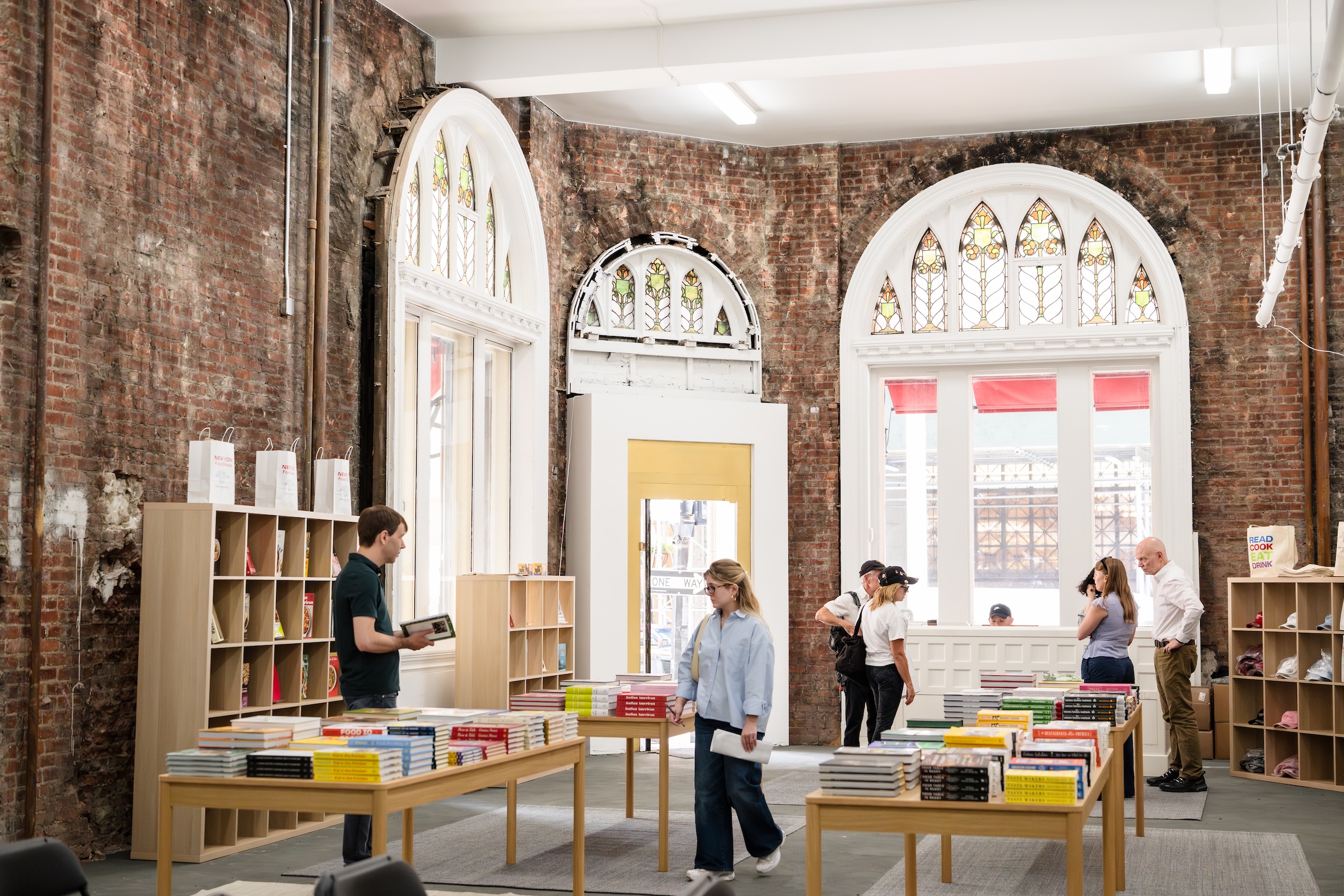 A lively bookstore set inside a historic building with exposed red brick walls. Several light wood display tables are arranged across the room, stacked with neatly organized books. Customers browse the selections, including a man flipping through a book on the left and a woman walking in a blue button-down shirt near the center. Light wood grid shelving units line the walls. The architectural highlights include large, white-trimmed arched windows and a central doorway, all topped with decorative stained-glass panels.
