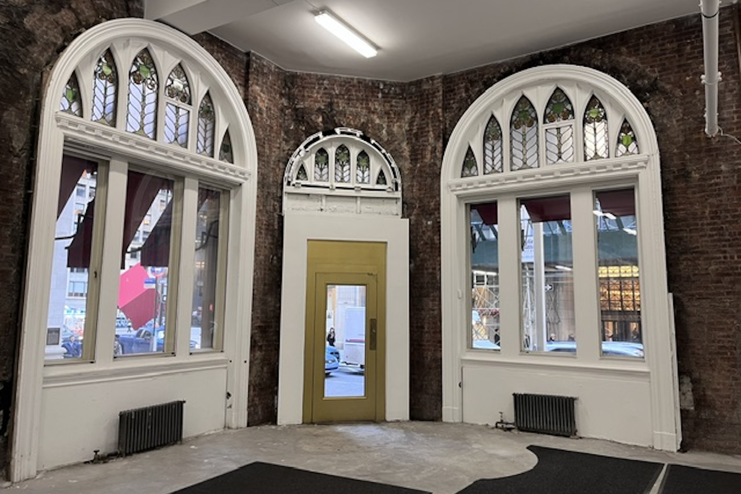 A vacant, historic commercial space with unfinished concrete floors and raw, exposed red brick walls. The focal point is a curved back wall featuring two large, white-trimmed arched window frames and a central doorway with a gold-colored frame, all under construction. The tops of the windows and door are fitted with detailed, arched stained-glass panels. Old iron radiator units sit along the baseboards, and a fluorescent tube light illuminates the empty room from above.