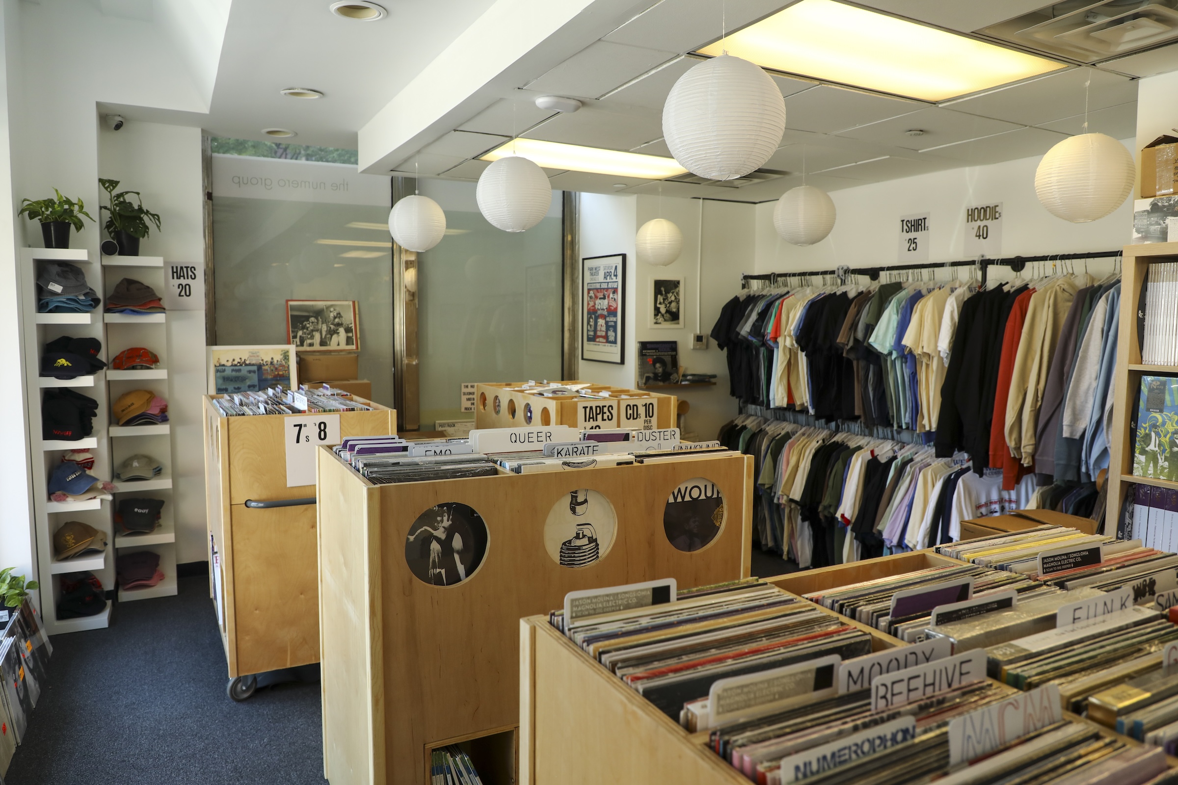 A vibrant, well-organized pop-up record and clothing store. Large, custom wooden bins filled with vinyl records dominate the foreground and center, labeled with genre tags like "QUEER," "KARATE," and "DUSTER." Spherical white paper lantern lamps hang from the ceiling. The right wall features double-tiered clothing racks packed with t-shirts and hoodies, while the left wall displays a tall white shelving unit stacked with baseball caps.