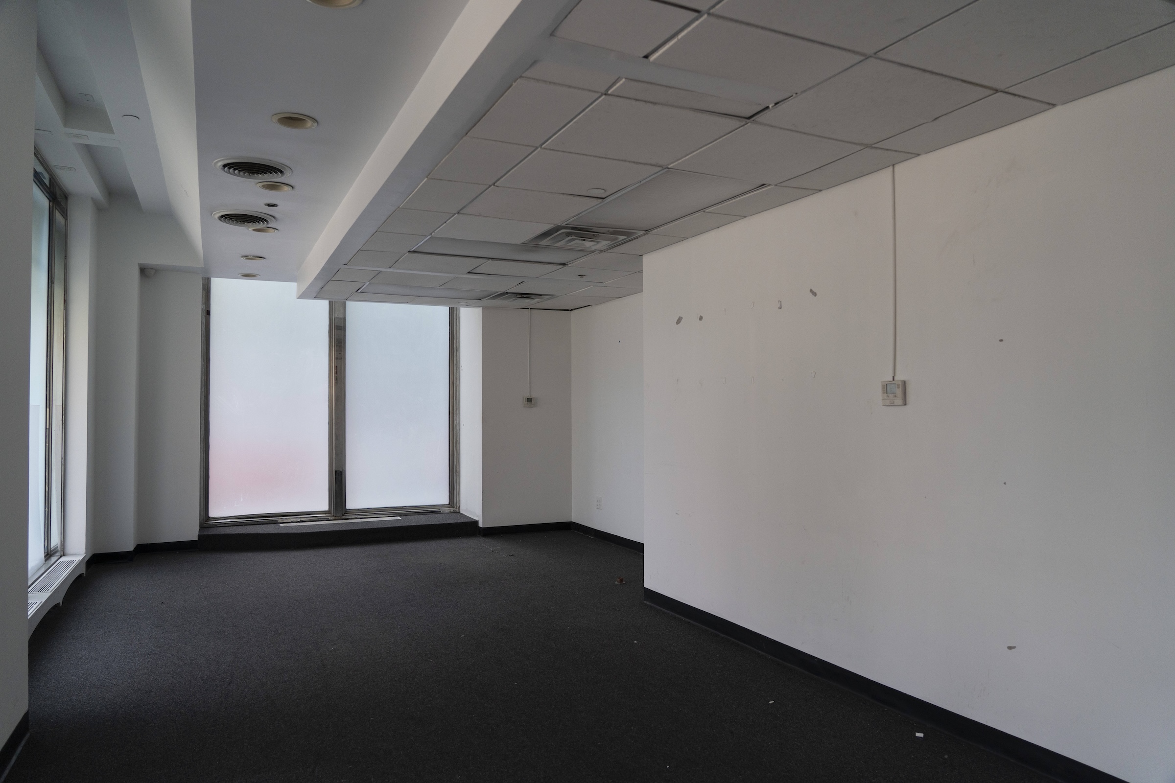 A completely vacant, stark commercial room with dark charcoal gray carpeting and scuffed white walls. The dropped ceiling features standard grid tiles, recessed lighting, and circular ventilation vents. A frosted glass window wall stands at the far end of the narrow room, casting soft, diffused daylight across the empty space.