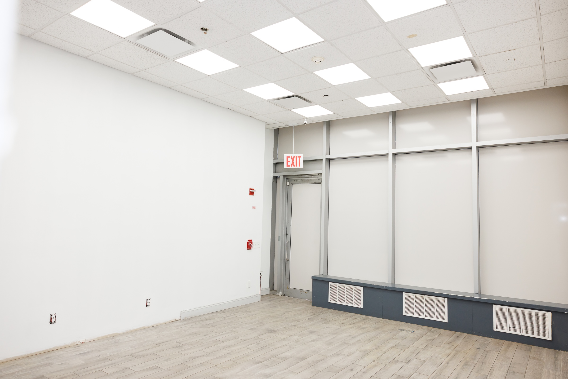 An empty retail room with light wood-plank flooring and bare white walls. The grid ceiling is fitted with square fluorescent light panels. At the back of the room, a glass partition wall and door feature frosted privacy film, with an illuminated red "EXIT" sign hanging above the doorway. A low, dark gray built-in bench with three white ventilation grates runs along the base of the glass window wall.