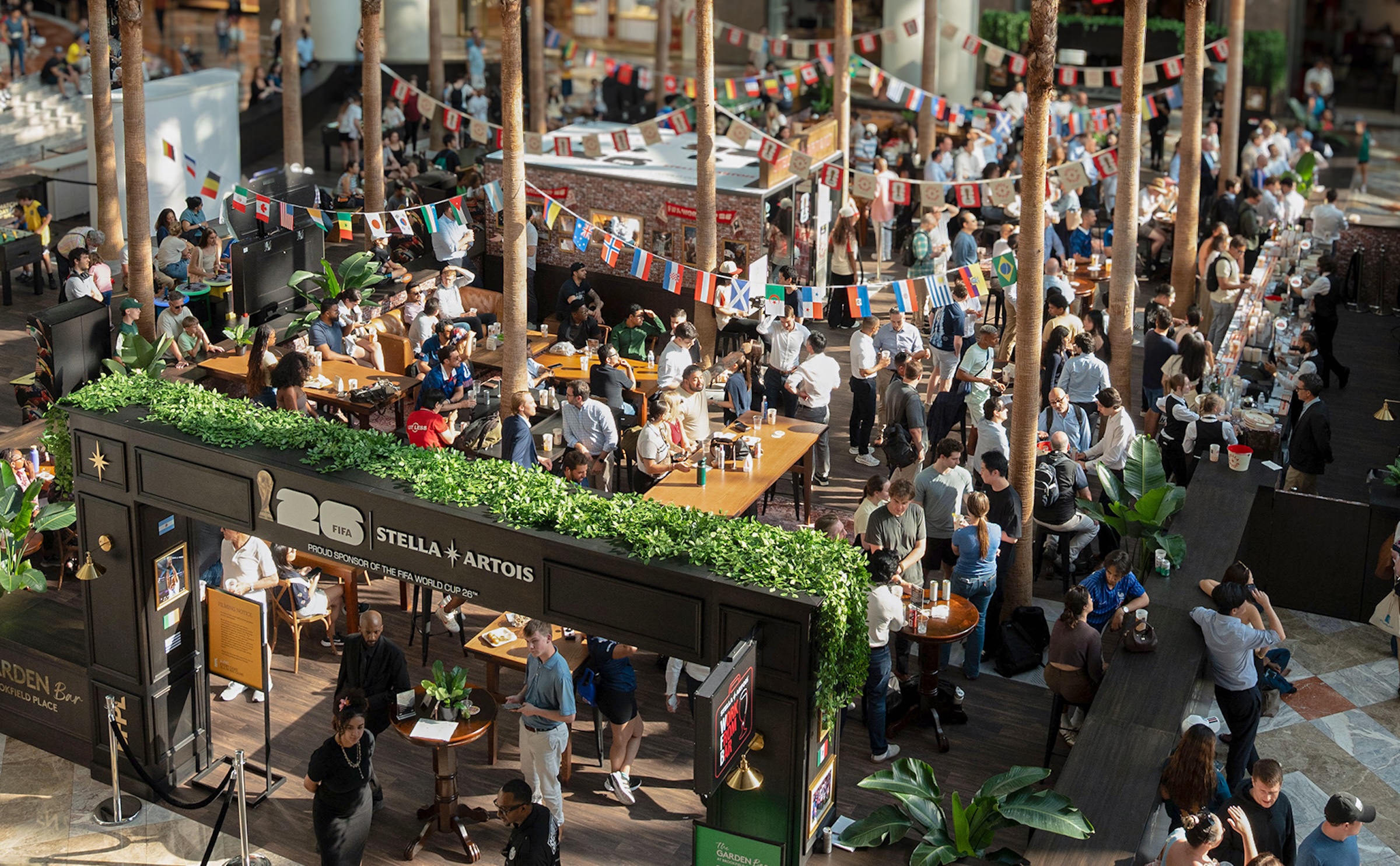 A high-angle, overhead shot of a large indoor gathering space packed with people. The venue resembles a courtyard or atrium, filled with rows of tall, slender palm trees stretching upward. Strung between the trees are numerous lines of small flags representing different countries. In the foreground, a black structures with green hedges on top displays the text "26 FIFA STELLA ARTOIS PROUD SPONSOR OF THE FIFA WORLD CUP 26™". Patrons are seen socializing, standing, and sitting at wooden tables and long bars throughout the lively event space.