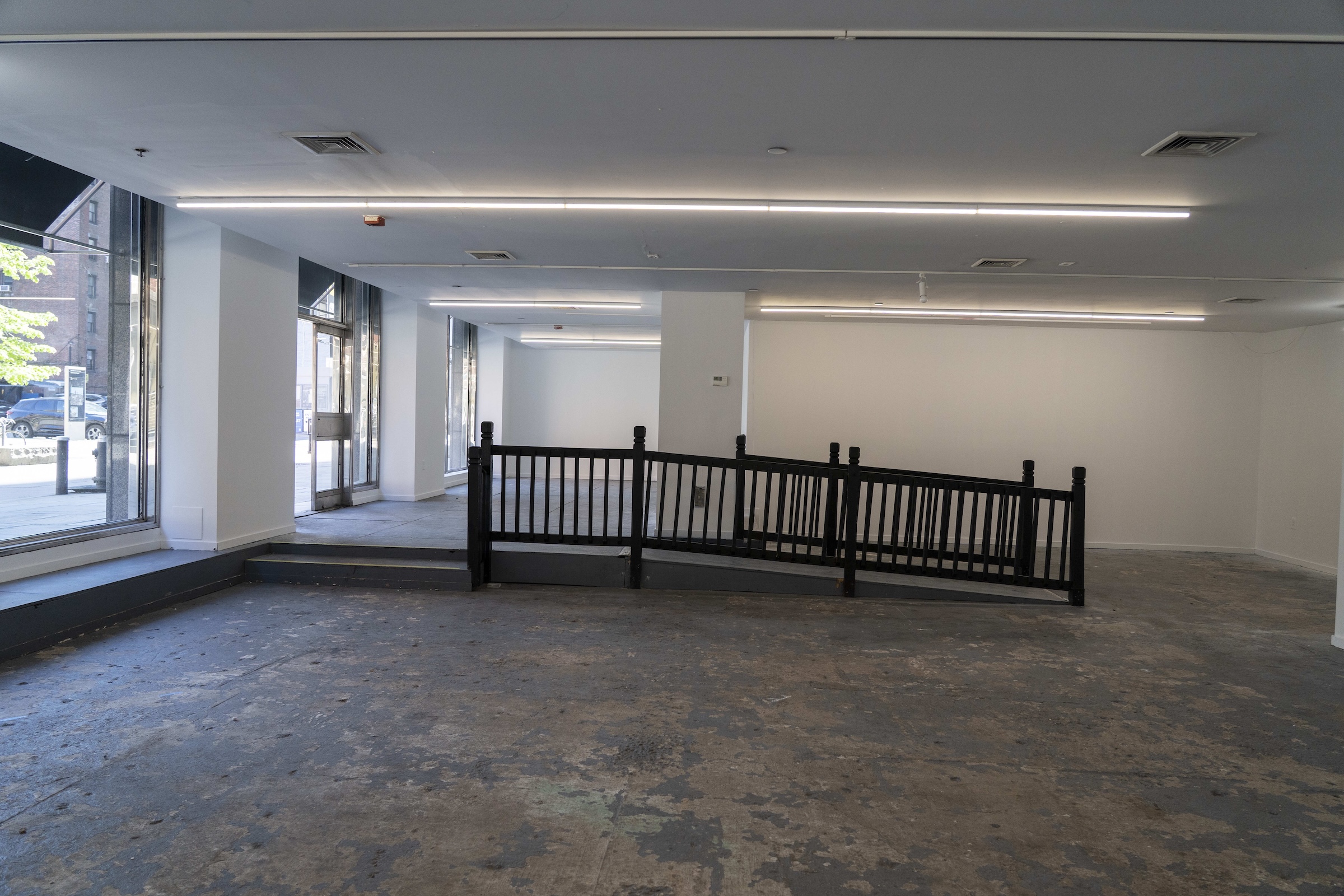 A wide shot of a spacious, completely empty commercial retail or gallery space with bare, worn concrete flooring and white walls. In the center of the room, a short dark wood or black wooden ramp with a matching vertical slat railing connects two slightly different floor levels. Long, thin fluorescent strip lights are mounted across the white ceiling. Large glass windows and an open doorway on the left look out onto a city street with a glimpse of parked vehicles and a building outside.
