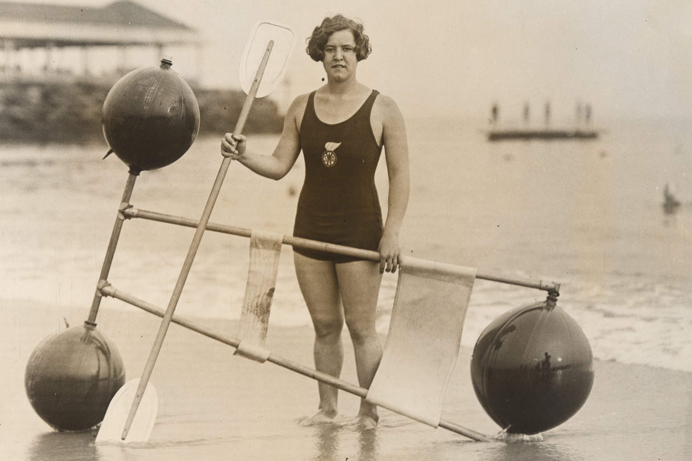 A vintage black-and-white photograph shows American swimmer Gertrude Ederle standing barefoot on a sandy beach at the water's edge. She wears a dark, fitted one-piece tank swimsuit with a round logo emblem featuring a winged symbol and the letters "WSA" (Women's Swimming Association) on her chest. Beside her is a large, custom-built mechanical water float contraption composed of a frame made from wooden or metal rods. The frame is attached to three large, dark spherical buoys—two resting near the ground and one raised on a vertical post—along with two oars or paddles and fabric slings suspended from the crossbars. In the background, calm ocean water stretches toward the horizon, where faint silhouettes of people stand on a distant platform or pier.
