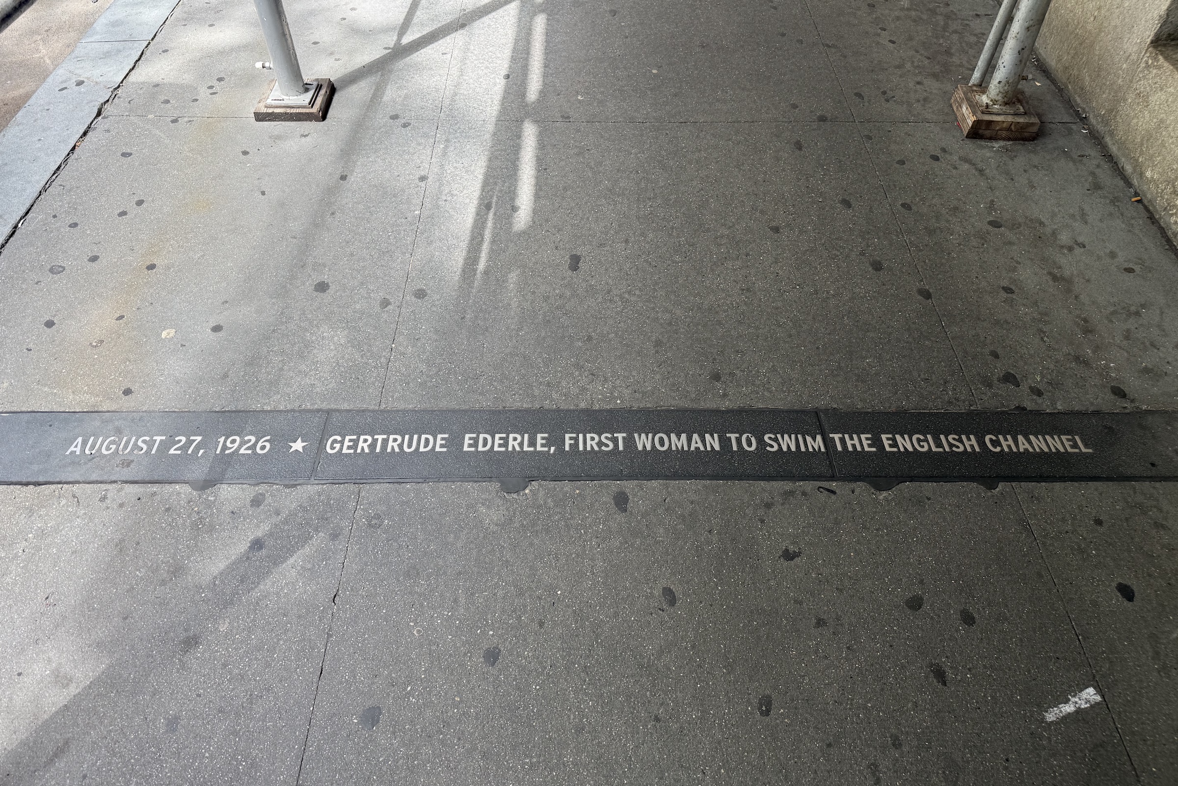 A wide shot looking down at a concrete city sidewalk featuring a dark granite strip embedded horizontally across the walkway. Engraved in white capital lettering on the dark strip is the inscription: "AUGUST 27, 1926 ★ GERTRUDE EDERLE, FIRST WOMAN TO SWIM THE ENGLISH CHANNEL". At the top of the frame, the bases of metal construction scaffolding poles rest on the gray pavement.