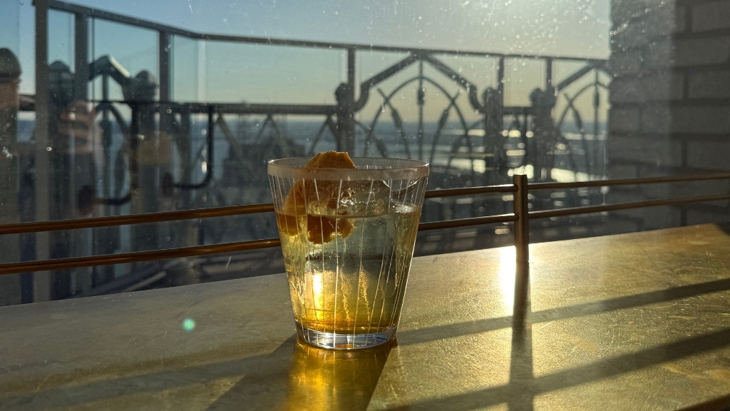 Short Alt-Text
A clear cocktail with a large ice cube and citrus twist sits on a sunlit wooden bar ledge overlooking a glass balcony and city skyline.

Detailed Alt-Text
A clear cocktail served over ice in a textured, ribbed glass sitting on a golden wooden counter bathed in warm, direct sunlight. Inside the glass, an orange citrus twist or garnish is visible against the ice. In the background, a glass barrier railing on an elevated balcony overlooks a distant, hazy skyline under a clear blue sky, casting long shadows across the bar top.