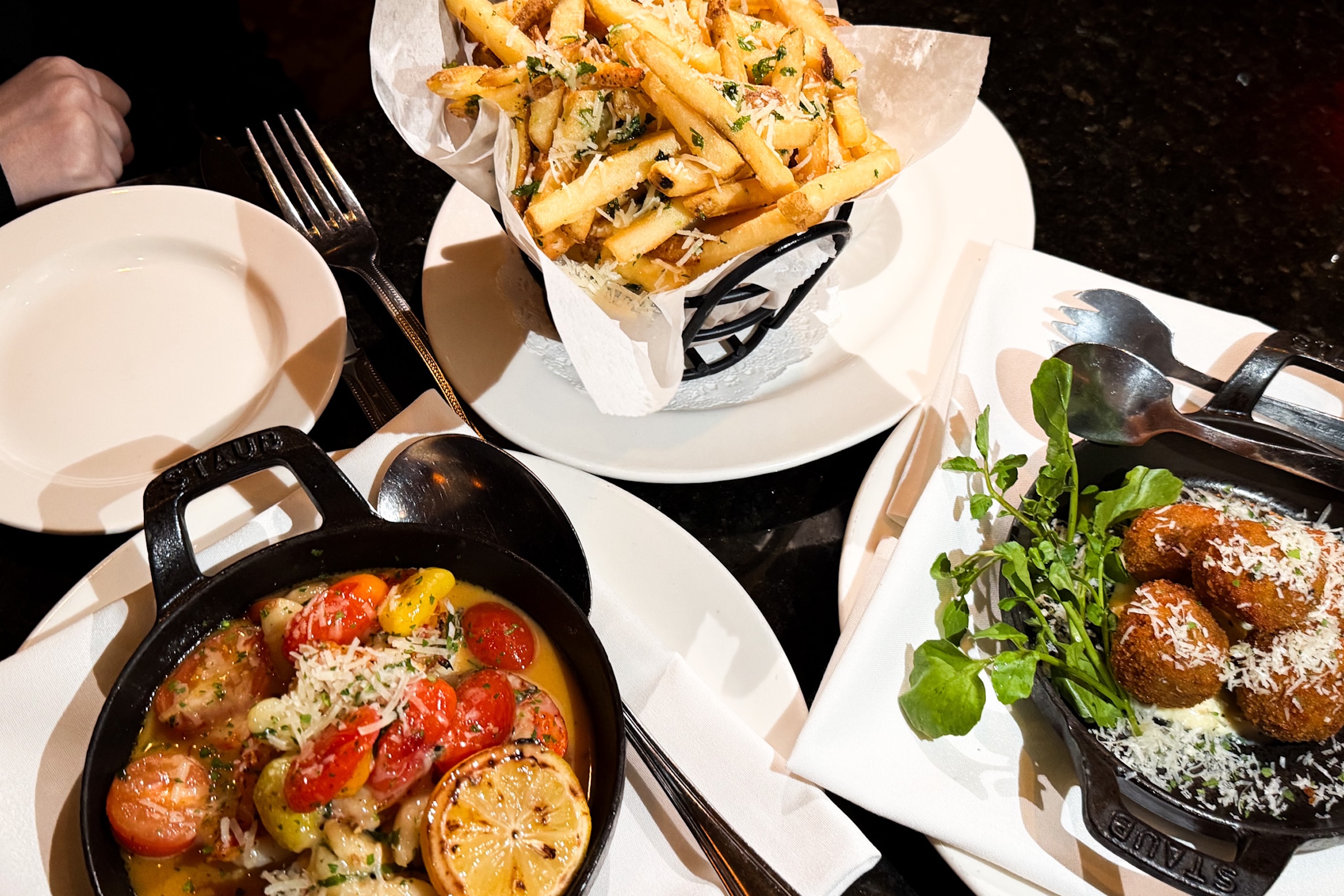 A top-down shot of a dark restaurant table spread with three dishes served in individual white plates. At the top center sits a wire cone lined with paper, filled high with french fries sprinkled with grated parmesan cheese and herbs. On the lower left, a black Staub cast-iron skillet holds a seafood dish with halved cherry tomatoes, herbs, melted butter, and a seared lemon wheel. On the lower right, another black cast-iron skillet contains fried croquettes topped with shredded cheese, garnished with fresh greens. An empty white side plate and silver utensils sit nearby.