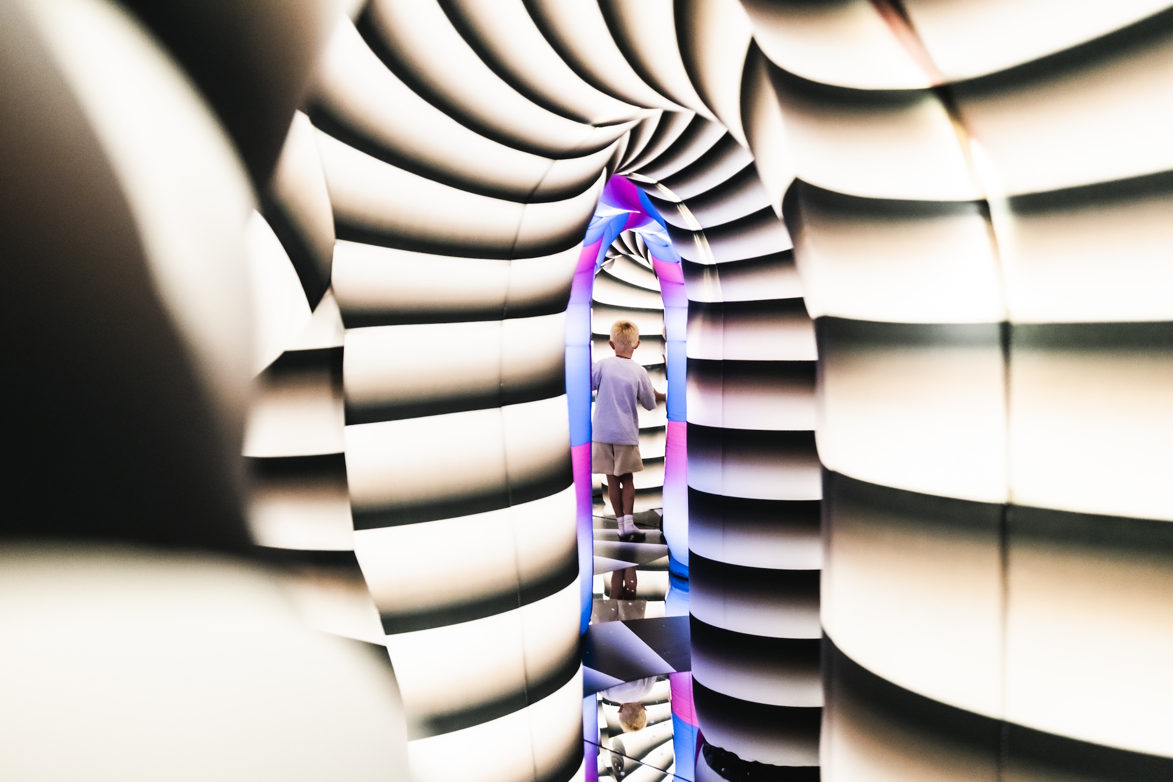 A young child standing inside a futuristic, spiraling tunnel with black-and-white striped inflatable walls, leading toward a brightly lit, neon pink and blue doorway.