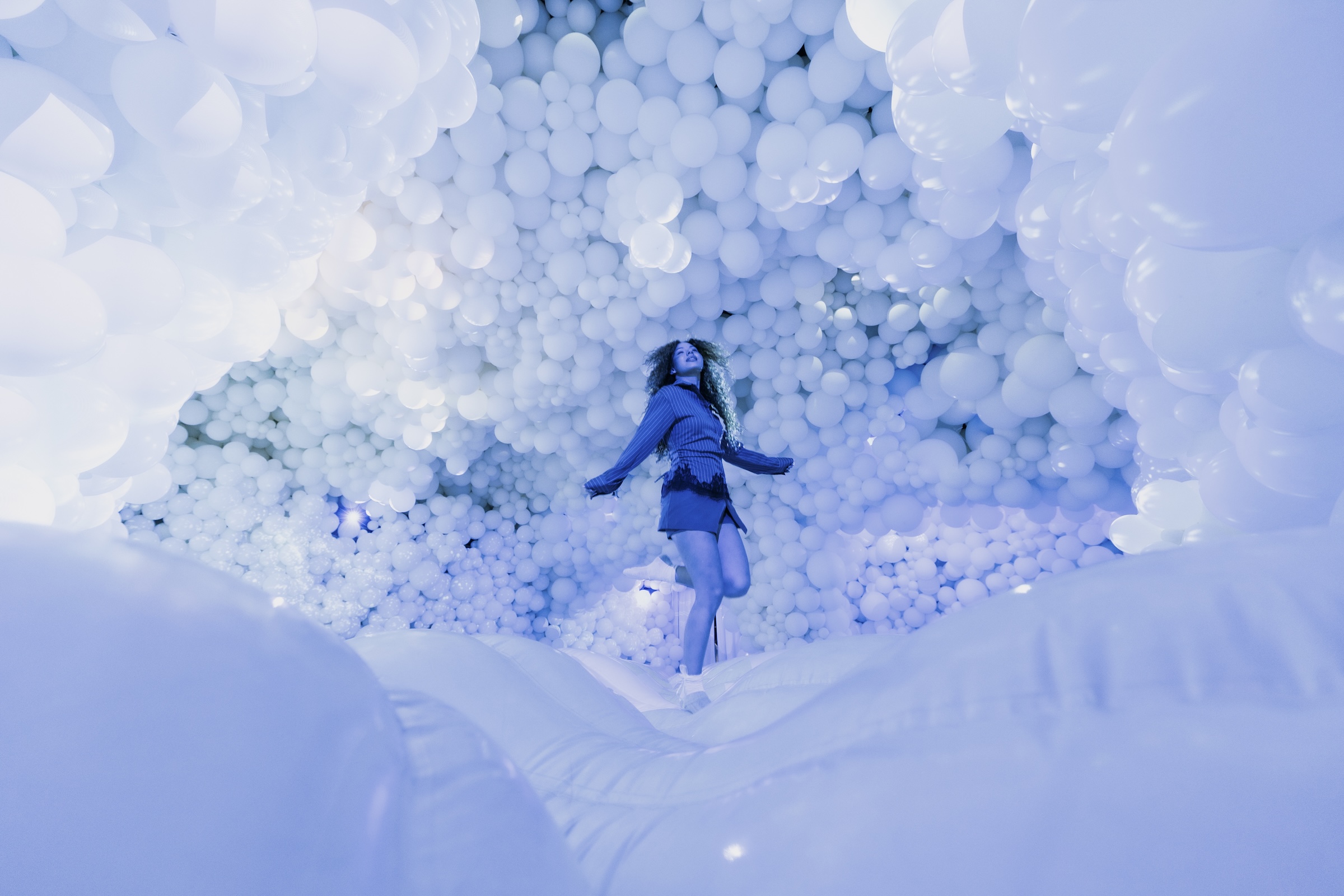 A person with curly hair wearing a blue outfit, captured mid-air as they jump on a white inflatable surface inside a room completely covered with white balloons bathed in cool blue lighting.