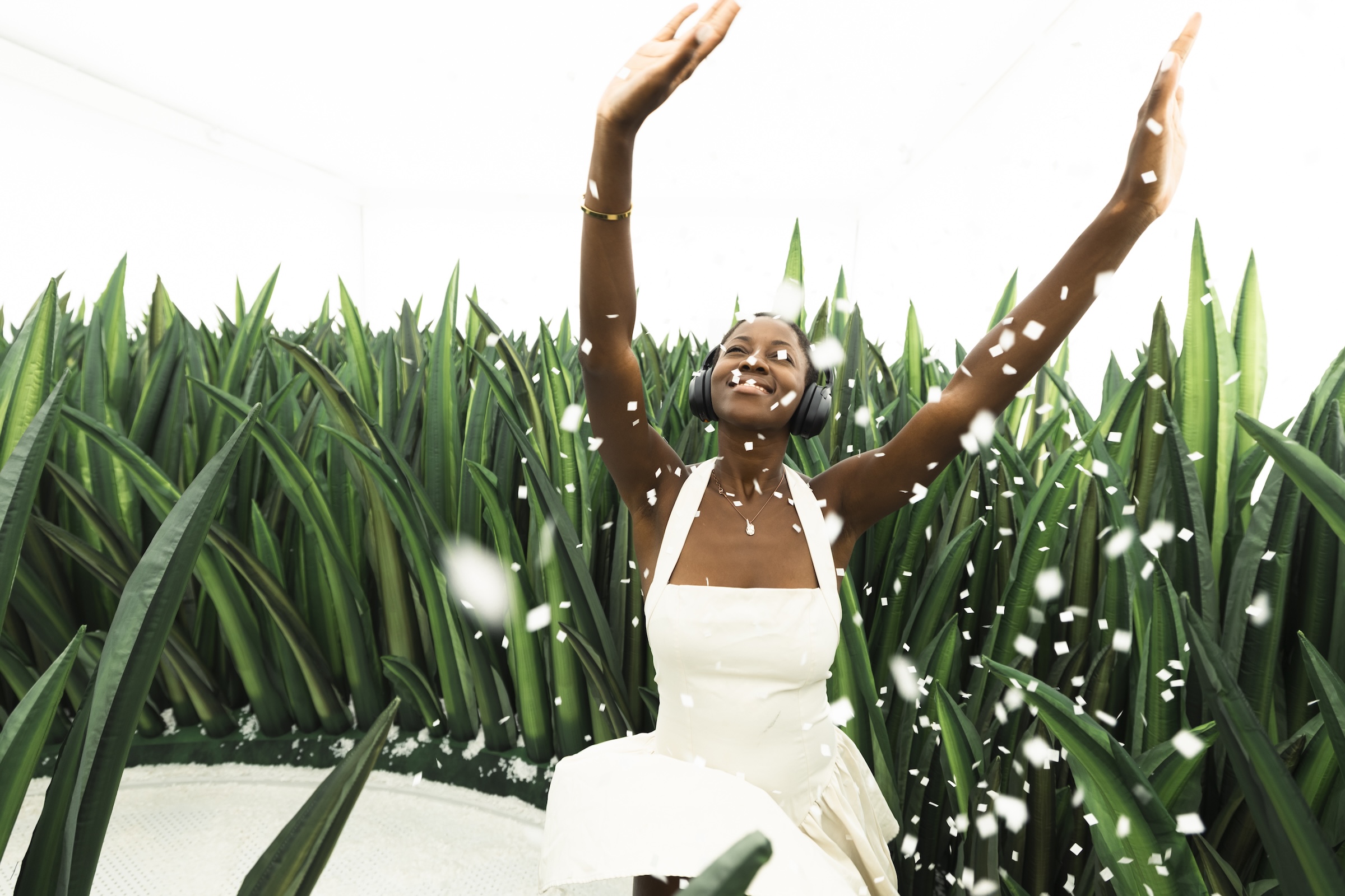 A smiling woman wearing a white dress and over-ear headphones, throwing white confetti into the air with both arms raised, surrounded by tall, spiky green plants in an artistic installation.