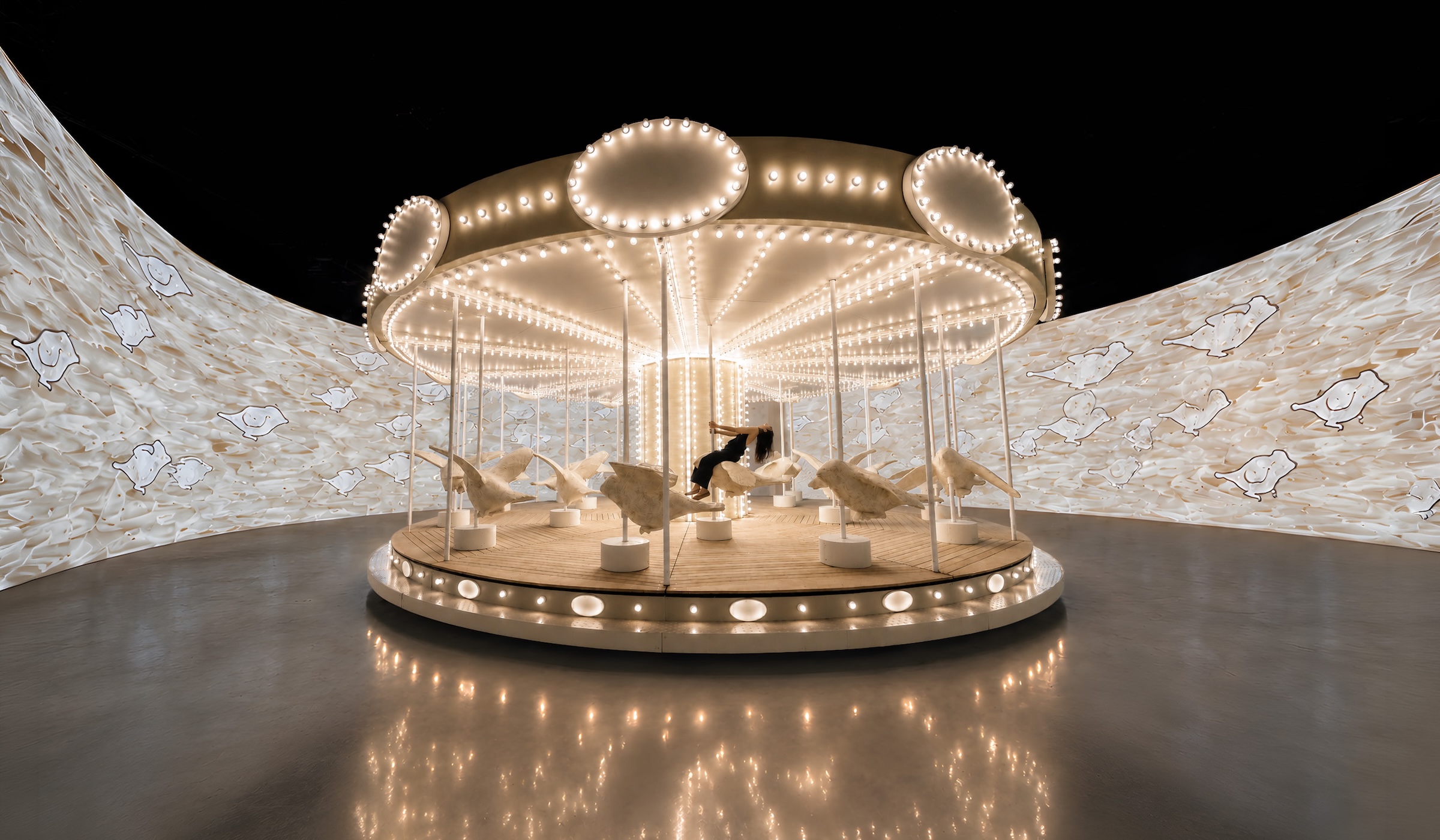 A brightly lit, vintage-style carousel featuring white bird sculptures inside a large room with curved walls displaying bird illustrations and a reflective dark floor.