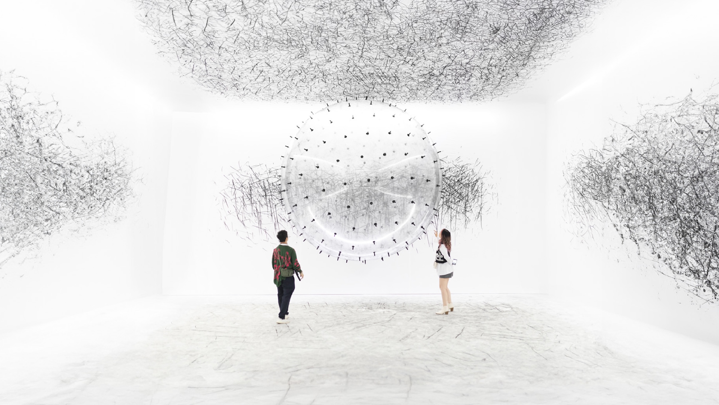Two visitors standing inside a stark white gallery space facing a giant, floating, spike-covered sphere that leaves charcoal scribbles across the surrounding white walls and ceiling.