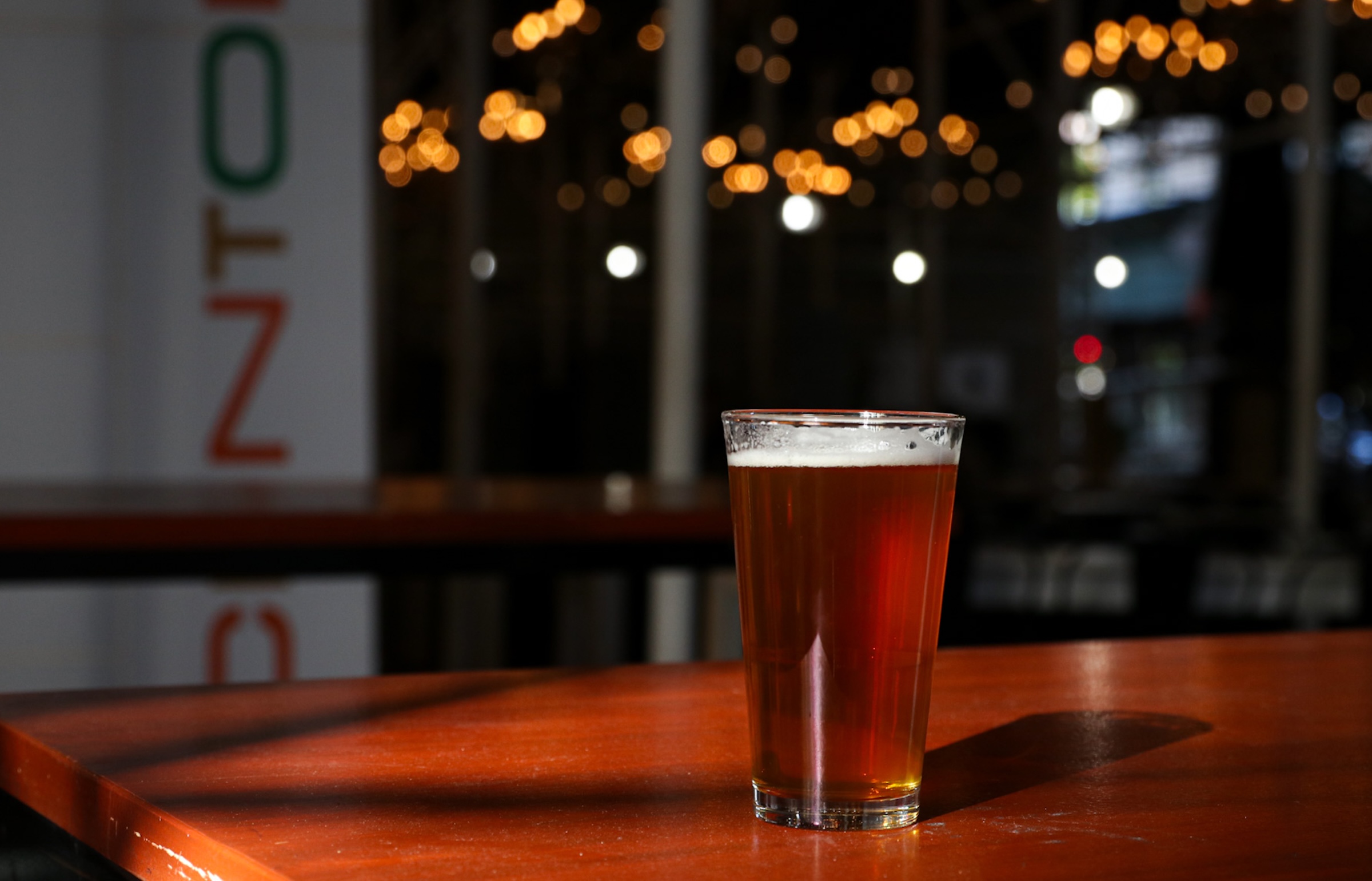 A pint glass filled with amber beer sits on a dark wooden tabletop. The background features blurred warm string lights and vertical lettering on a wall.