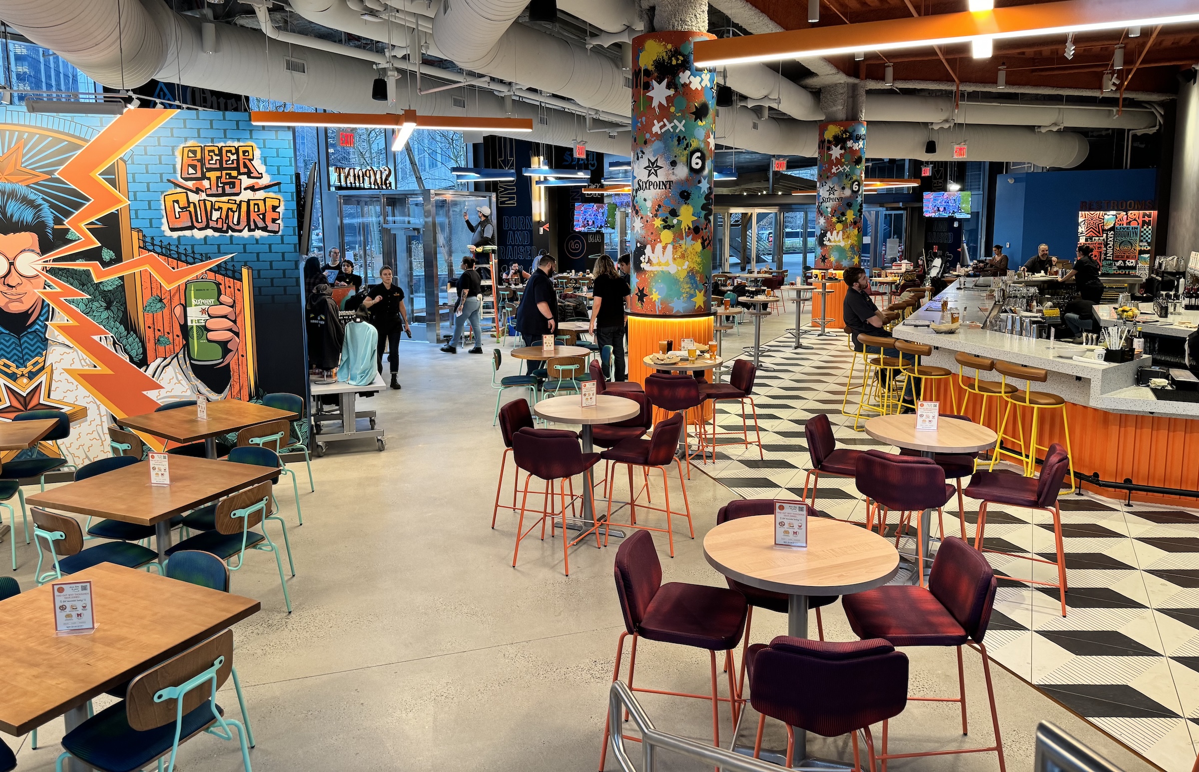 Spacious indoor taproom featuring colorful pop-art wall murals and decorated structural pillars. The seating area includes high-top tables with maroon chairs, wooden tables with teal chairs, and a long bar with yellow barstools along a black-and-white patterned floor.