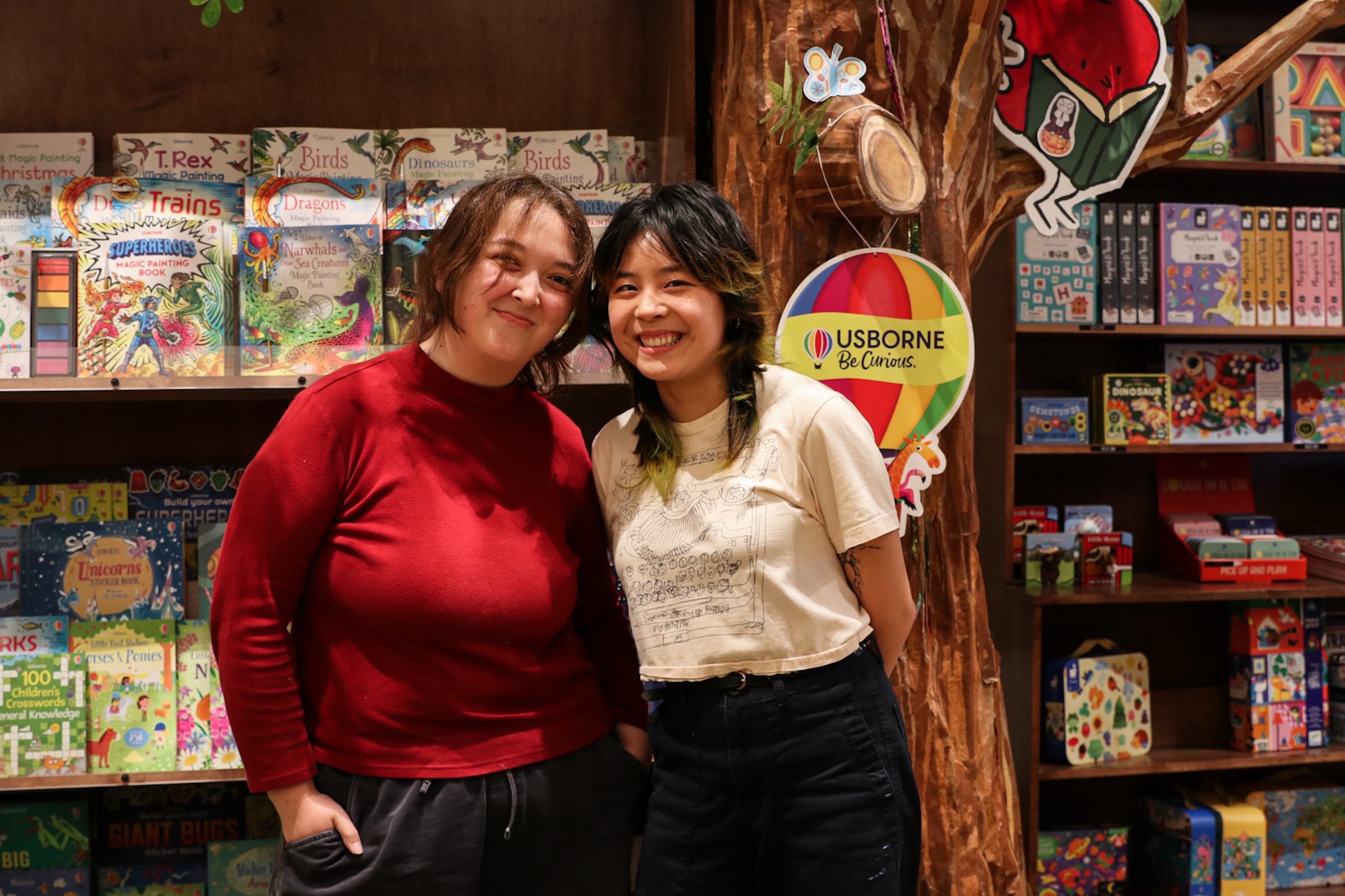 Two smiling women stand shoulder-to-shoulder inside a bookstore, posing next to a decorative paper-mache tree structure and shelves filled with colorful children's books and activities.