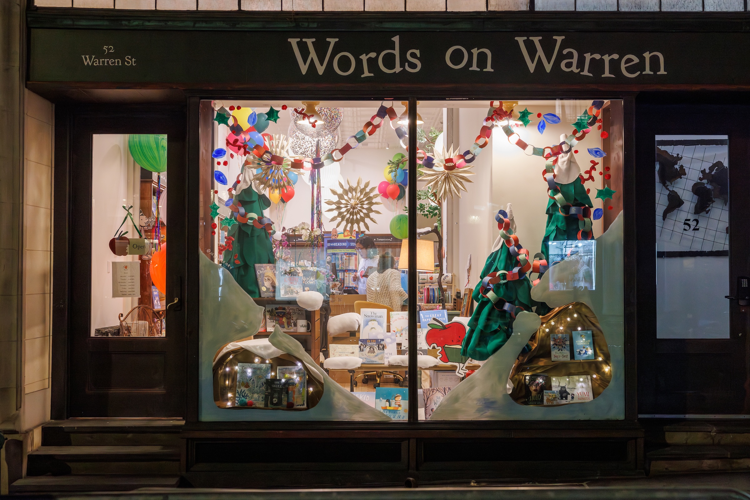 A nighttime storefront view of "Words on Warren," featuring brightly lit display windows filled with festive decorations, including paper chain garlands, geometric stars, balloon clusters, stylized fabric Christmas trees, and children's books arranged throughout the display.